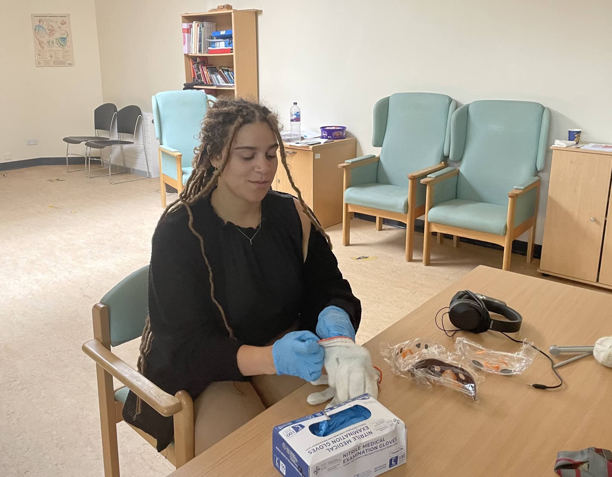 Person with braided hair sitting at a table, wearing blue gloves, with safety items like goggles and masks on the table. - Home Instead