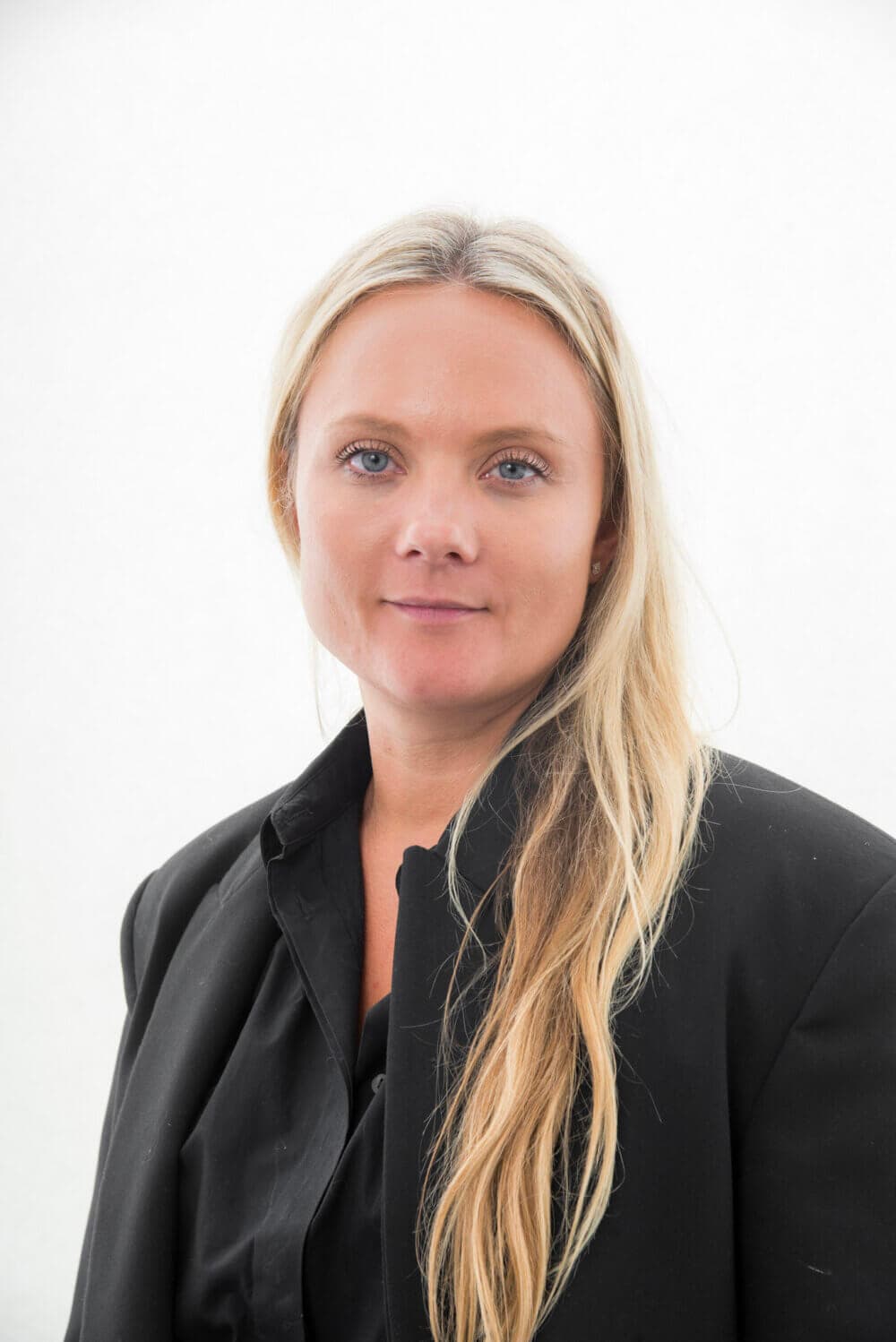 A person with long blonde hair wearing a black blazer and black top, standing against a plain white background. - Home Instead