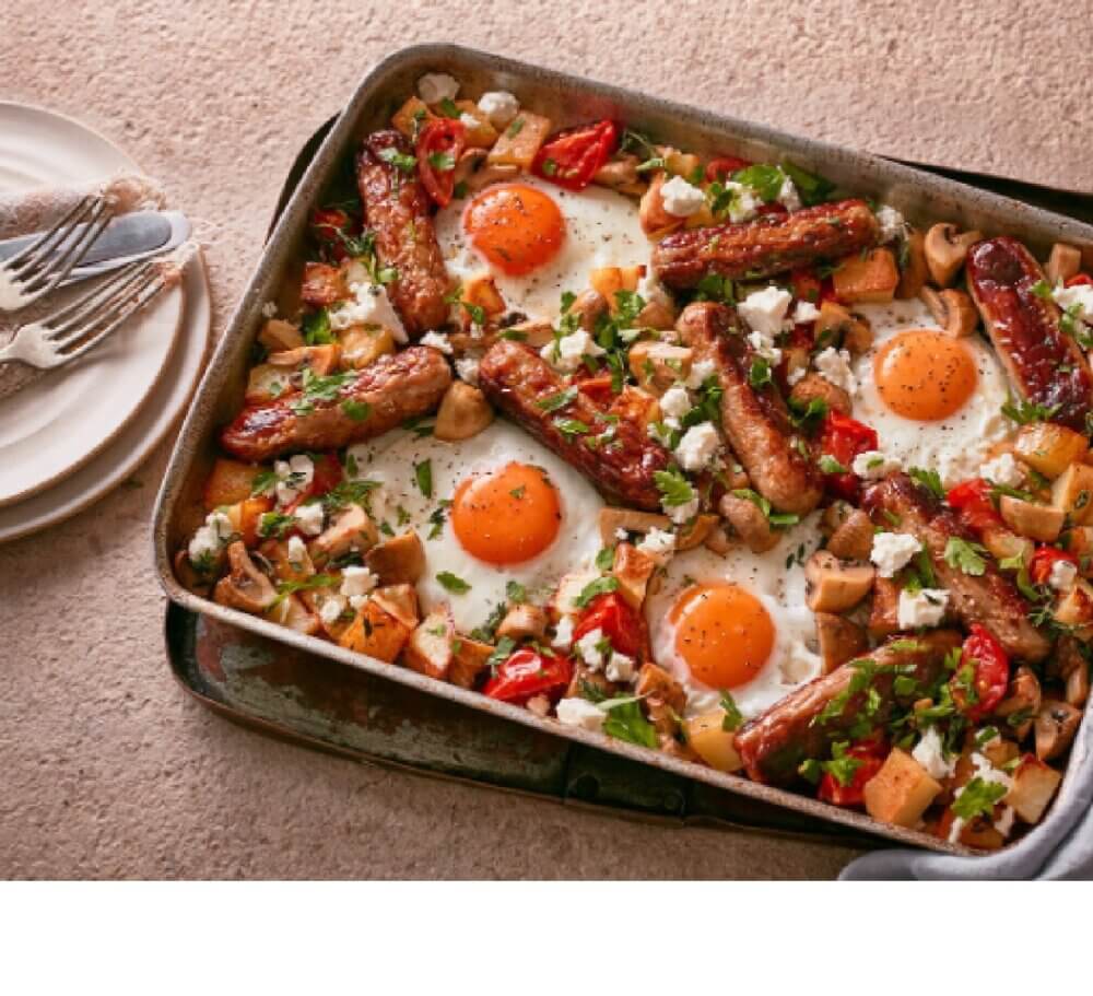 Baked eggs with sausages, cherry tomatoes, mushrooms, and herbs on a baking tray, next to a stack of plates and utensils. - Home Instead