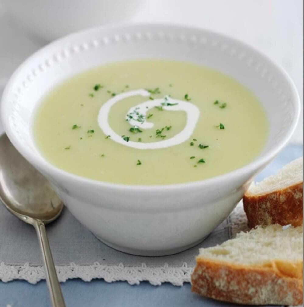 A bowl of creamy soup garnished with herbs and a swirl of cream, served with slices of bread and a spoon on the side. - Home Instead