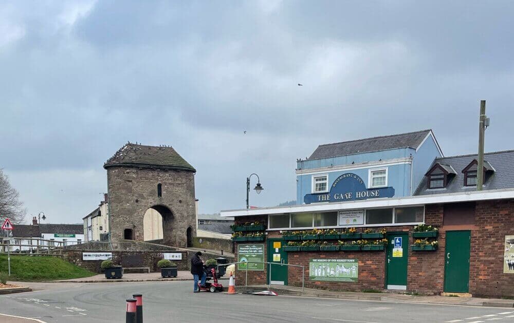 Historic brick gatehouse with arched entrance near a modern building. A person on a mobility scooter passes by. - Home Instead