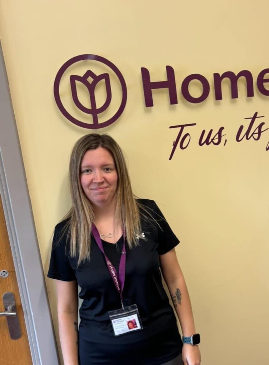 A woman in a black shirt stands in front of a wall with a logo and text, wearing a lanyard with an ID badge. - Home Instead
