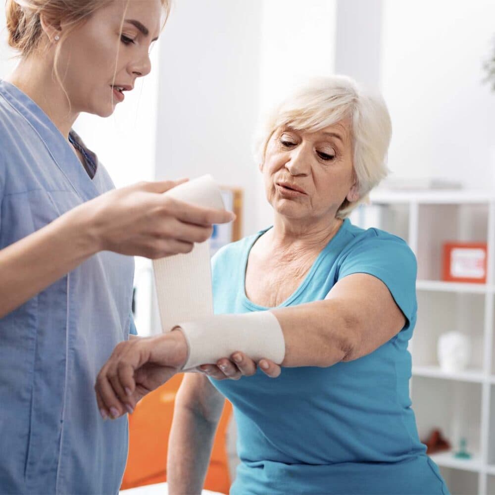 A nurse bandages an elderly woman's arm in a medical setting. The woman appears to be explaining something.
