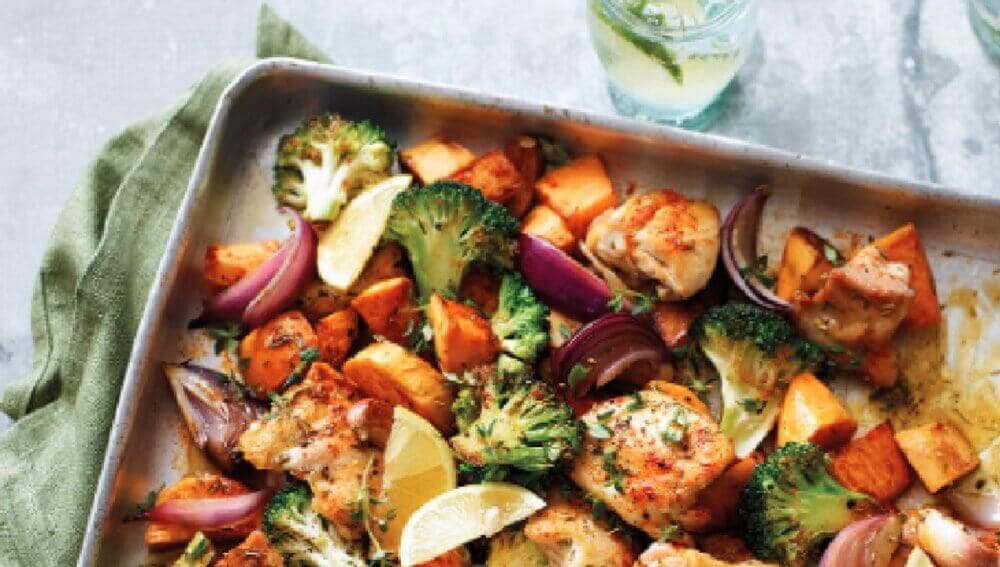 Roasted chicken, broccoli, sweet potatoes, and red onions on a sheet pan, garnished with lemon slices and herbs. - Home Instead