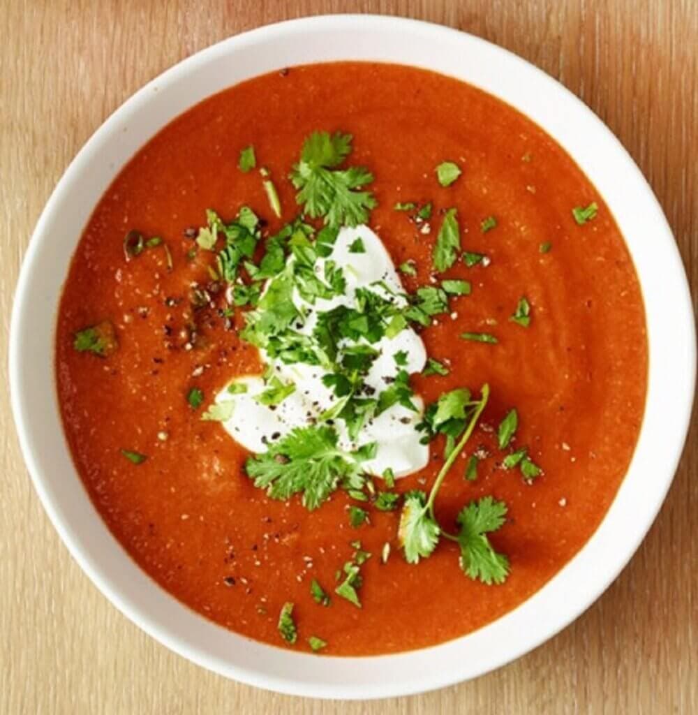 A white bowl filled with tomato soup garnished with a dollop of sour cream, chopped cilantro, and cracked black pepper. - Home Instead