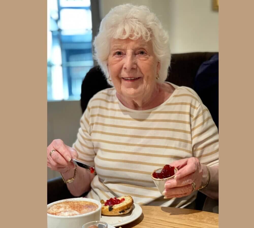 An elderly woman with white hair, smiling and enjoying a scone with jam and cream at a cafe. - Home Instead