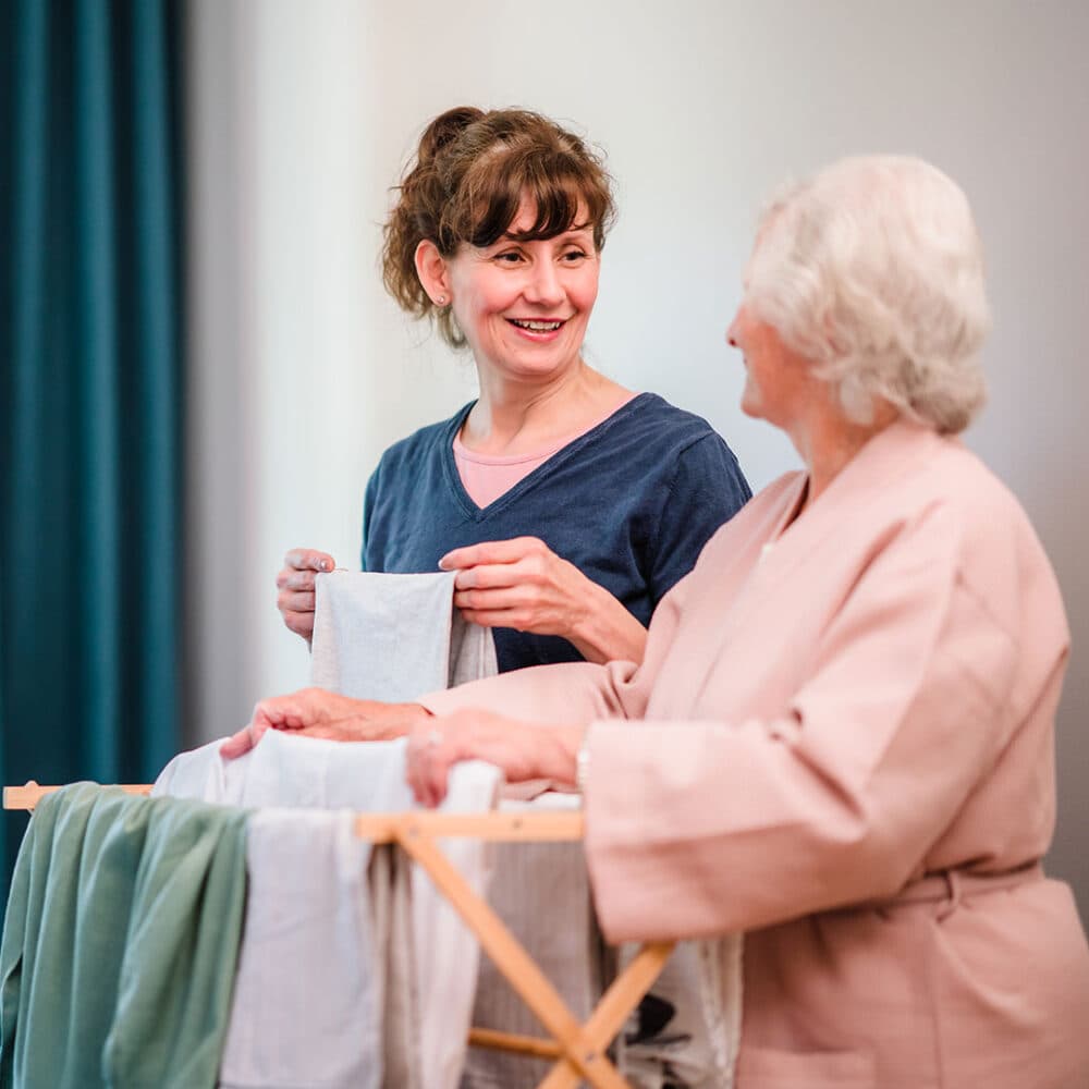 Two women smiling while folding laundry together, with clothes draped over a drying rack. - Home Instead