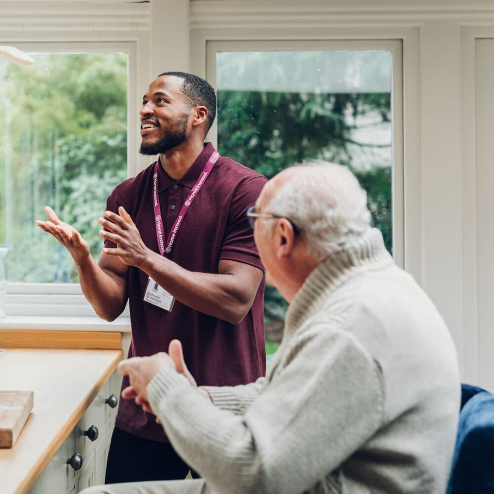 A caregiver wearing a maroon polo shirt talks and gestures to an elderly man in a gray sweater inside a sunlit room. - Home Instead