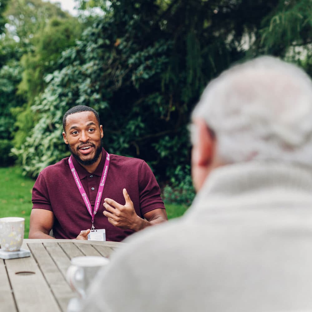 A man in a maroon shirt talks with an older person at an outdoor table, with trees in the background. - Home Instead