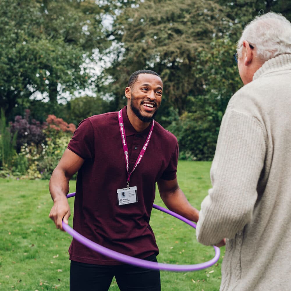 A man with a purple hula hoop engages with an older person in a garden setting. - Home Instead