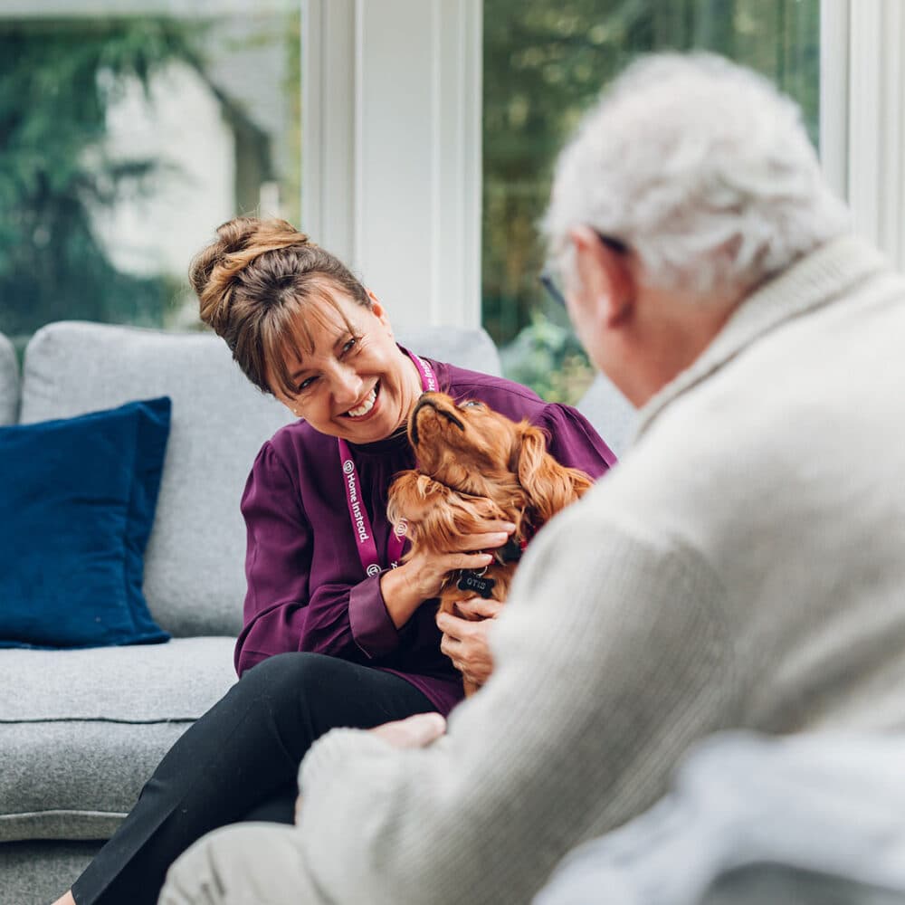 A woman sits next to an older man, smiling at a brown dog she is petting in a cozy living room. - Home Instead