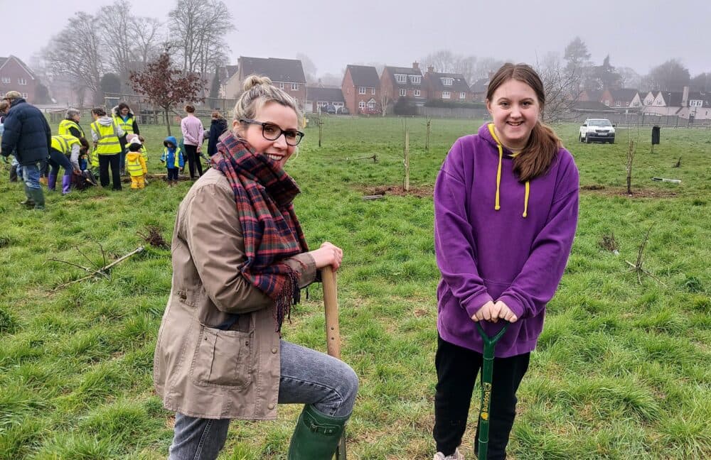 Two women in a park with shovels, participating in a tree planting event. People and houses in the background. - Home Instead