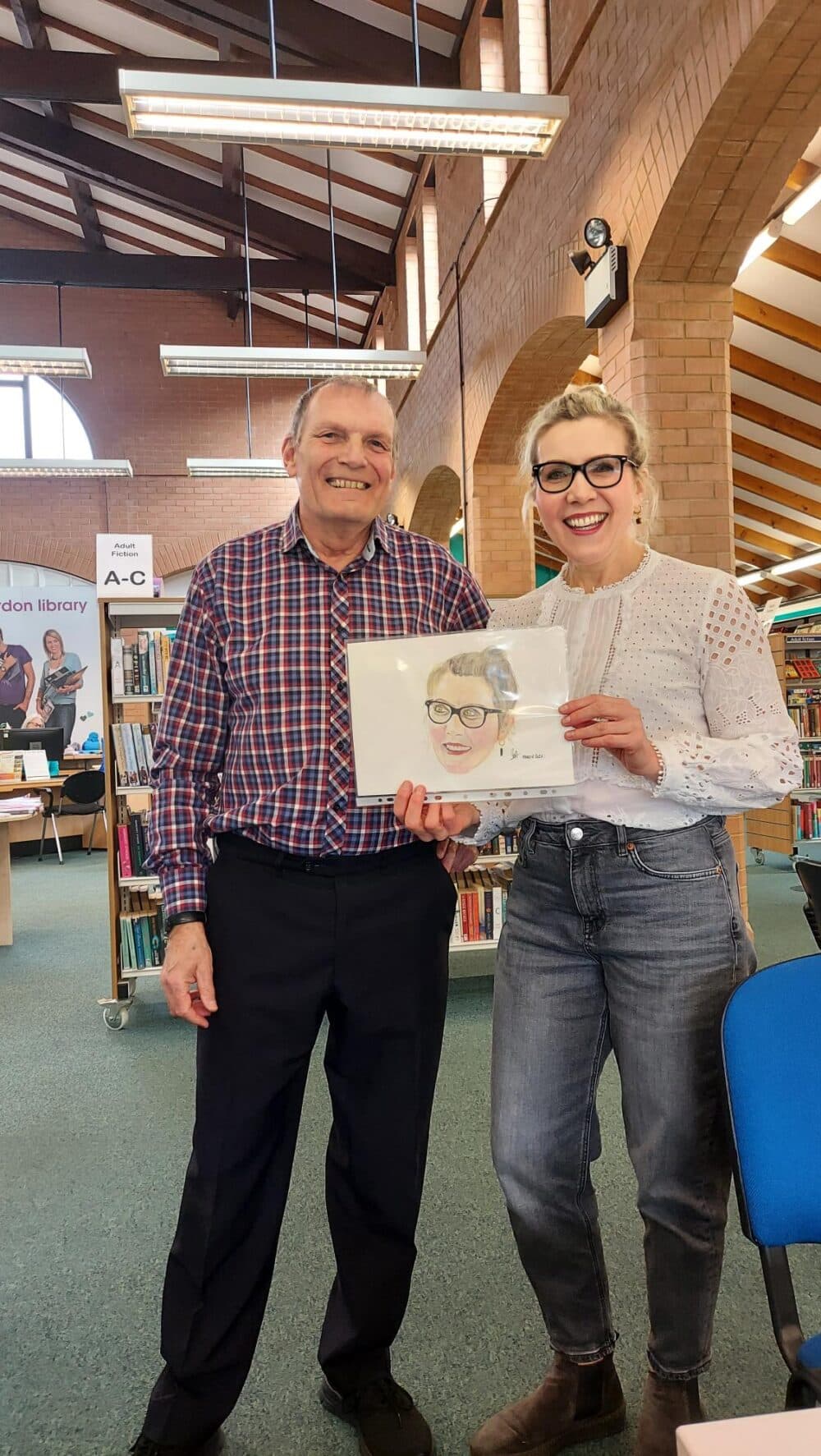 Two people smiling, one holding a drawing of a woman, in a library with bookshelves and an archway in the background. - Home Instead