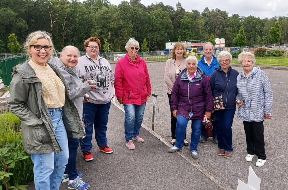 A group of nine smiling people standing outdoors on a paved area near a green park. - Home Instead