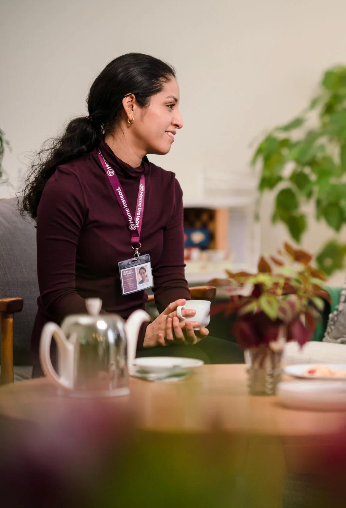 Woman with a lanyard and ID badge holding a cup seated at a table with a teapot and plants. - Home Instead