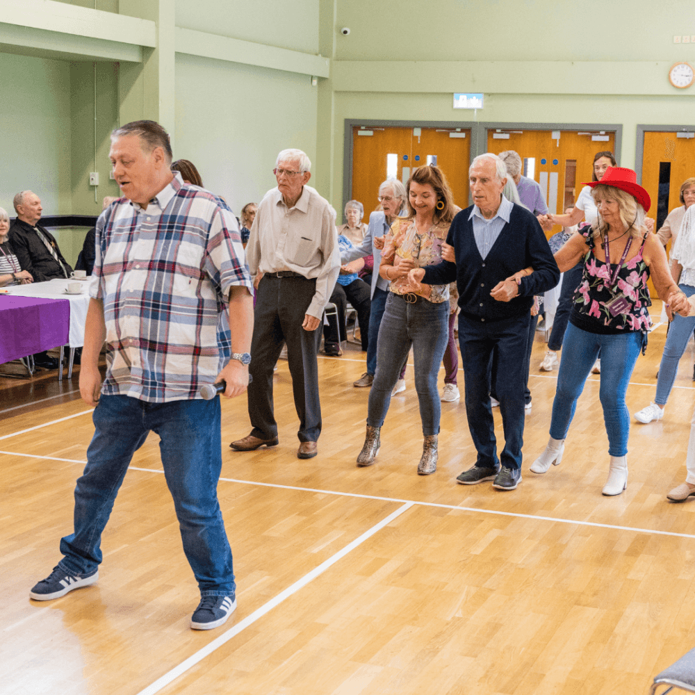 A group of people line dancing in a community hall with wooden floors and tables covered with purple and white tablecloths. - Home Instead