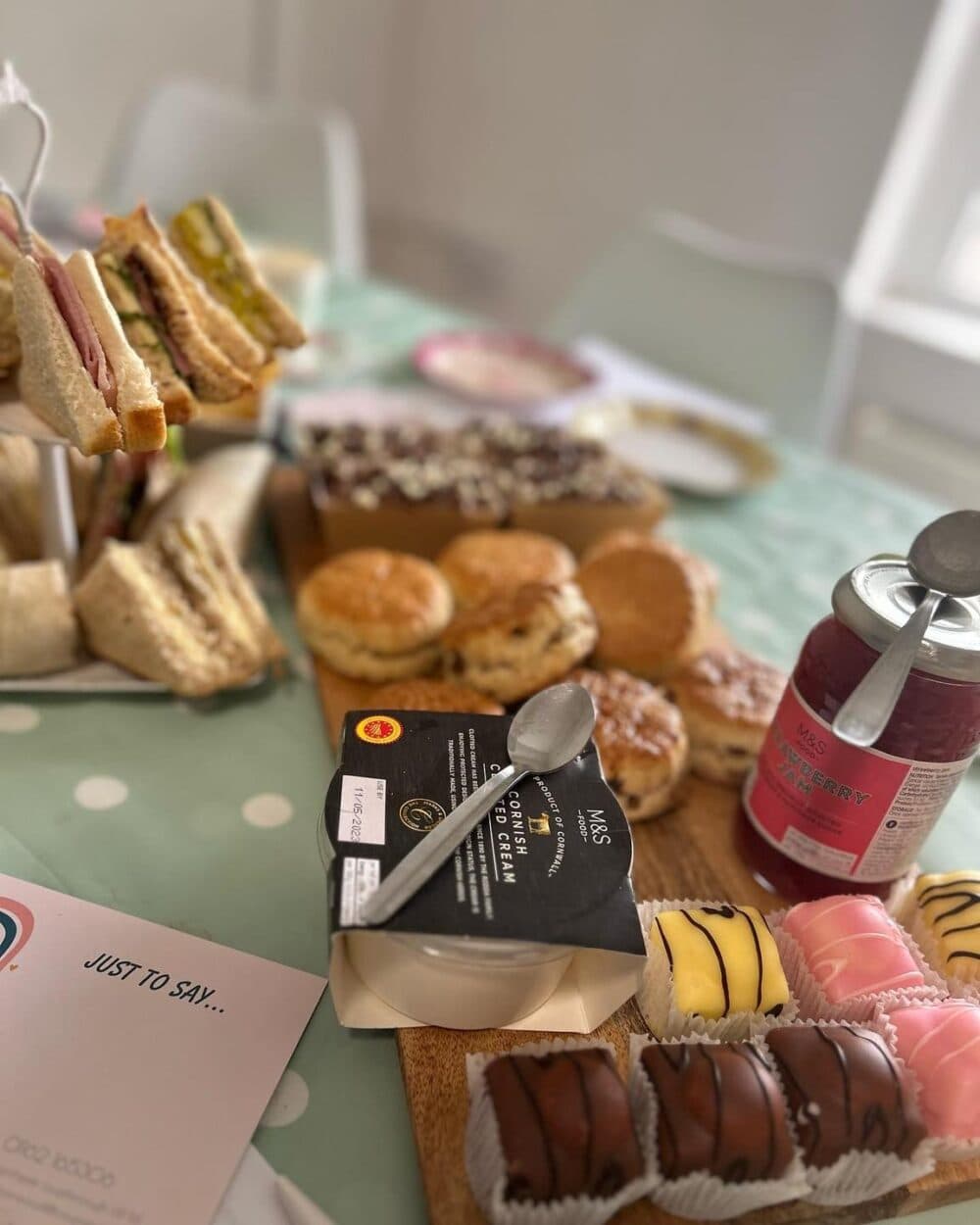 A table set for tea with sandwiches, scones, pastries, and a jar of raspberry preserve. - Home Instead