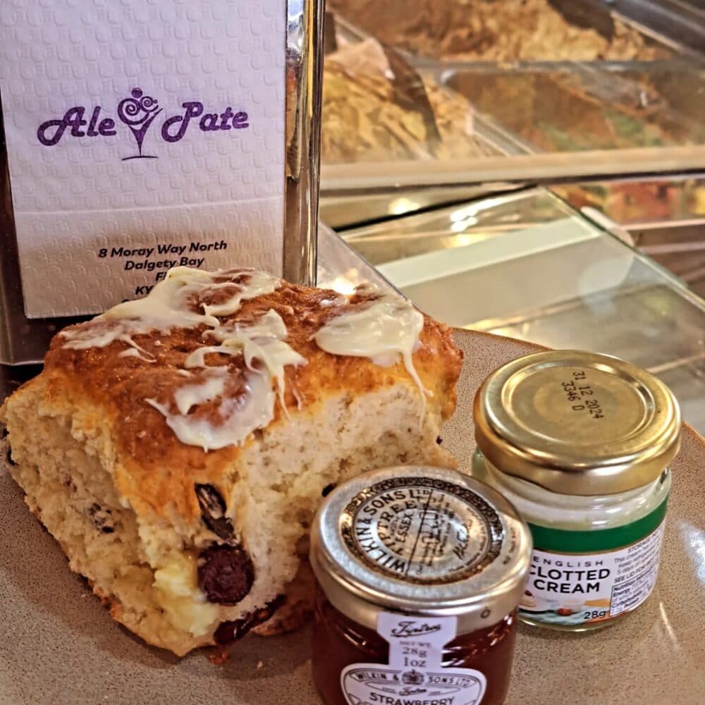 A scone with clotted cream and jam, served on a plate beside napkins from "Ale & Pate," with an array of pastries in the background. - Home Instead