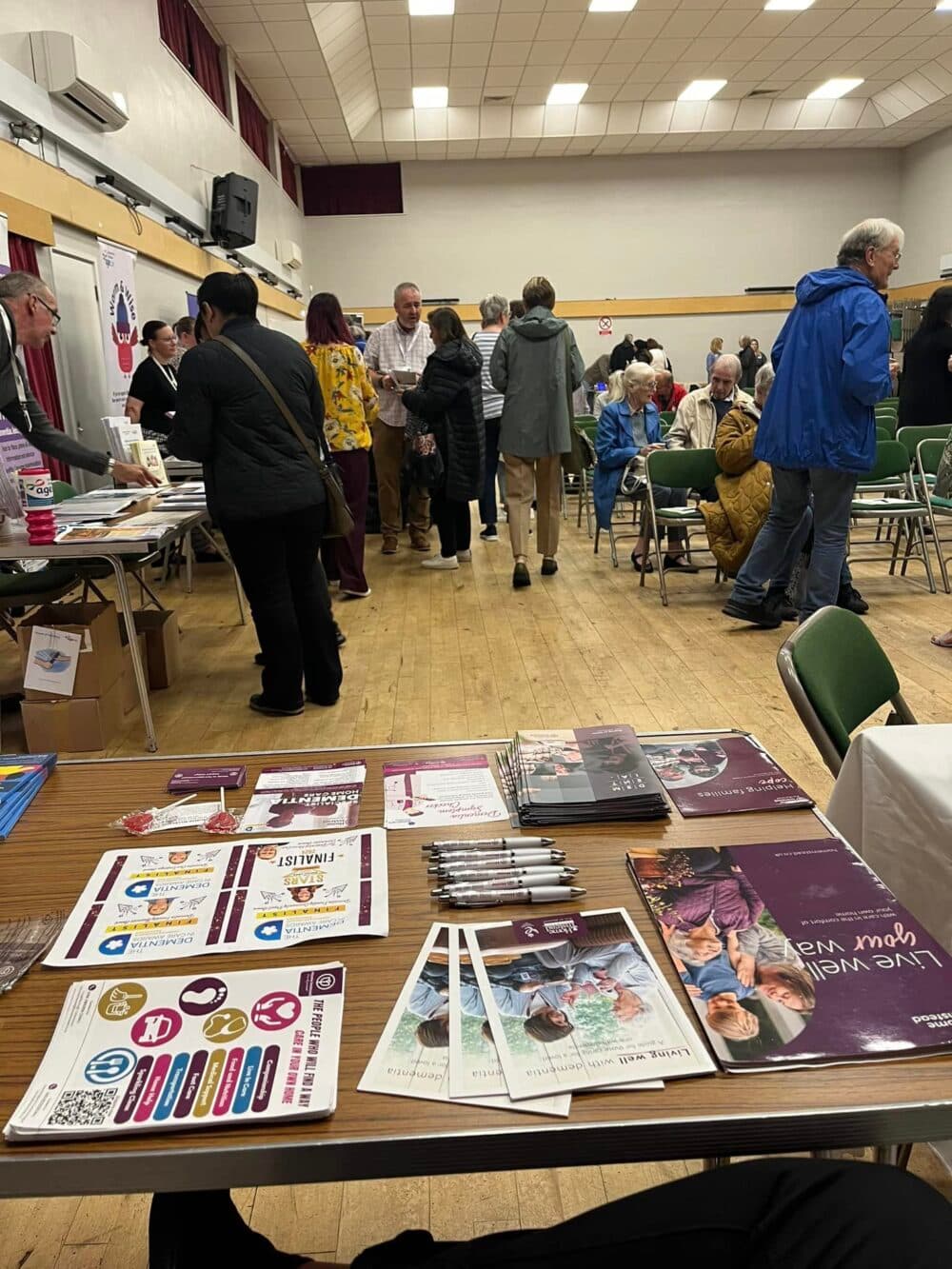 View from behind a table with pamphlets and pens at a busy community event in a hall with people interacting. - Home Instead