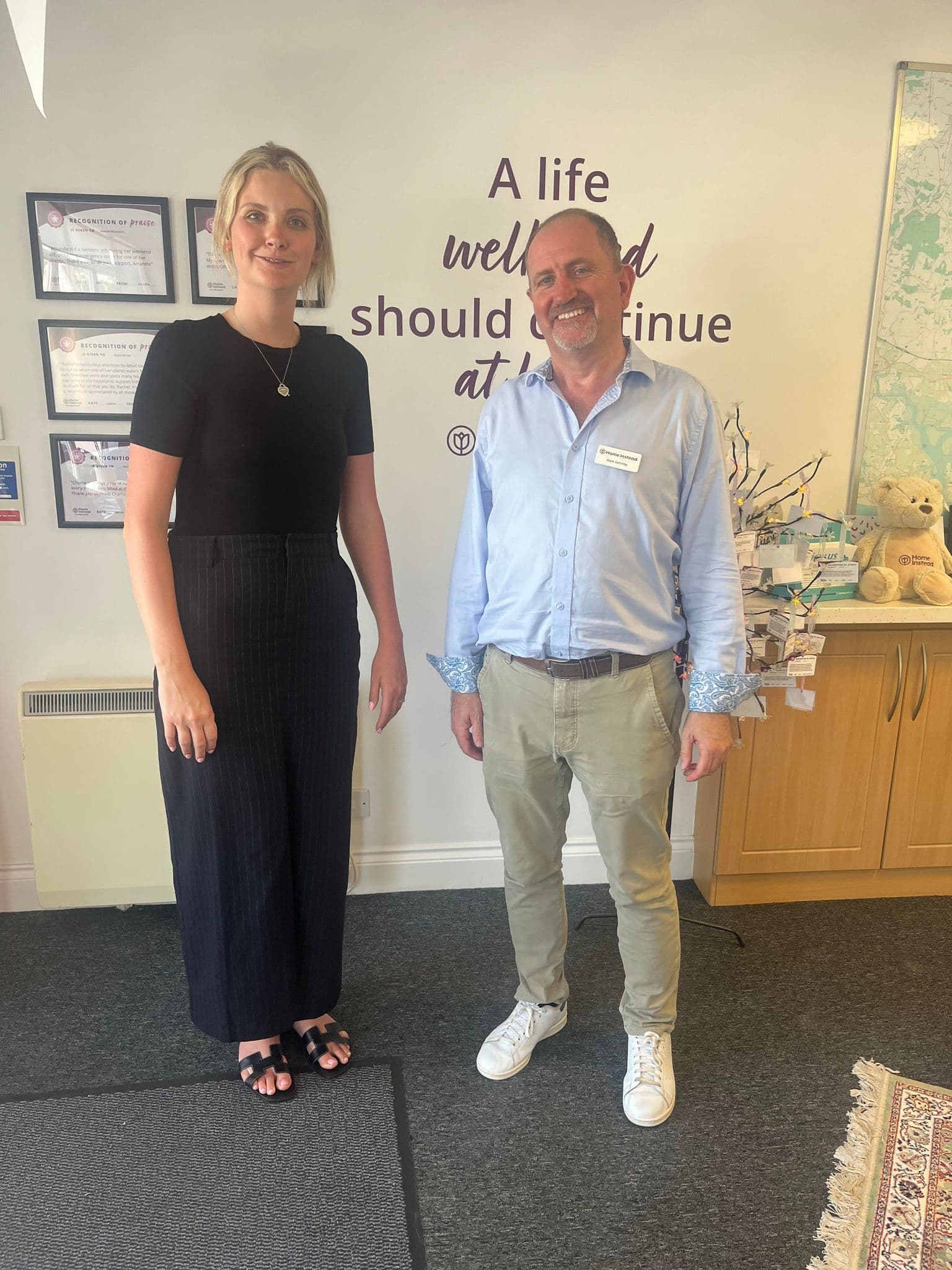Two people standing in an office, smiling at the camera. A wall quote and certificates are visible in the background. - Home Instead