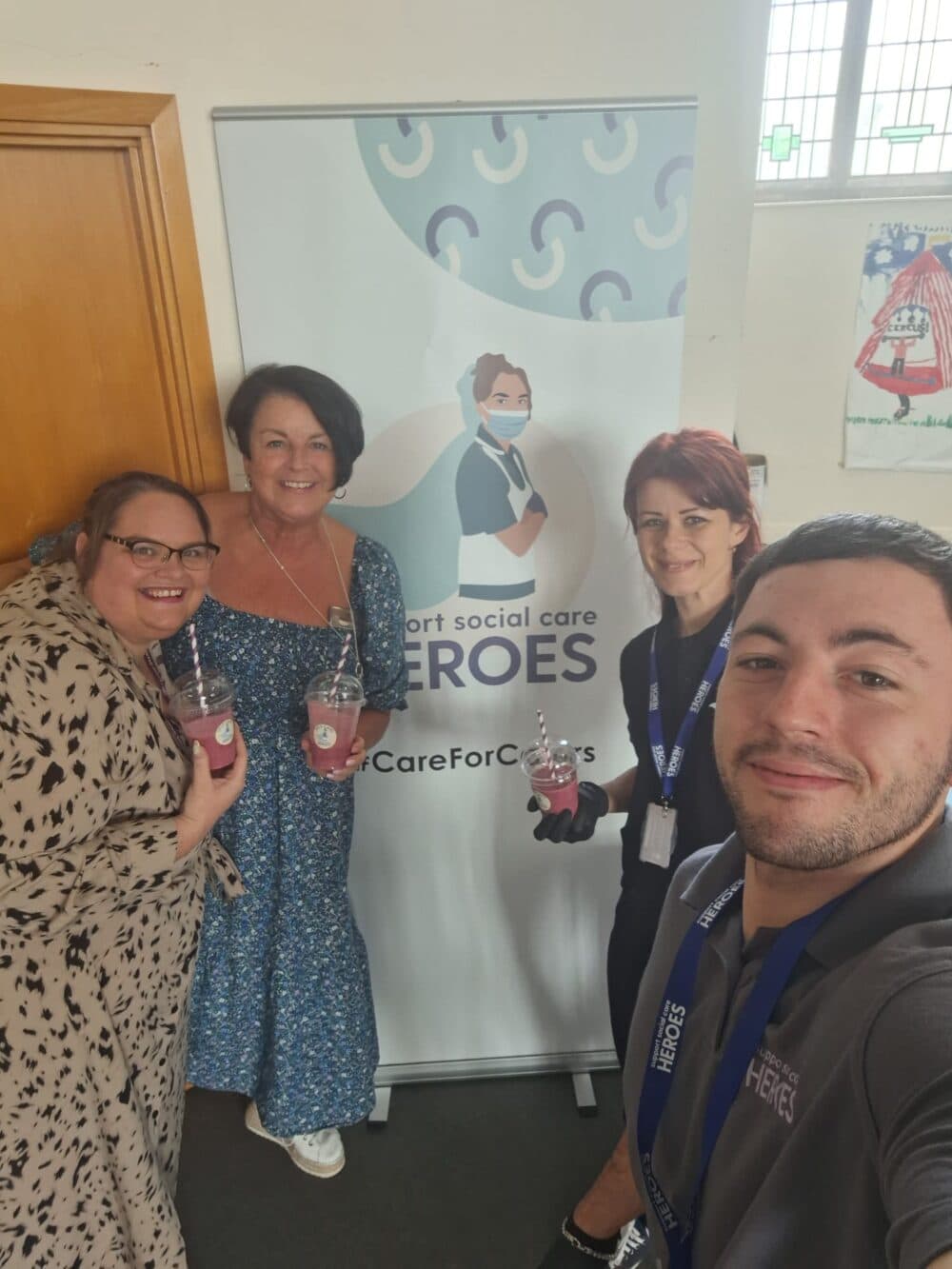 Four people smiling and holding drinks in front of a banner that says "Support Social Care Heroes. - Home Instead