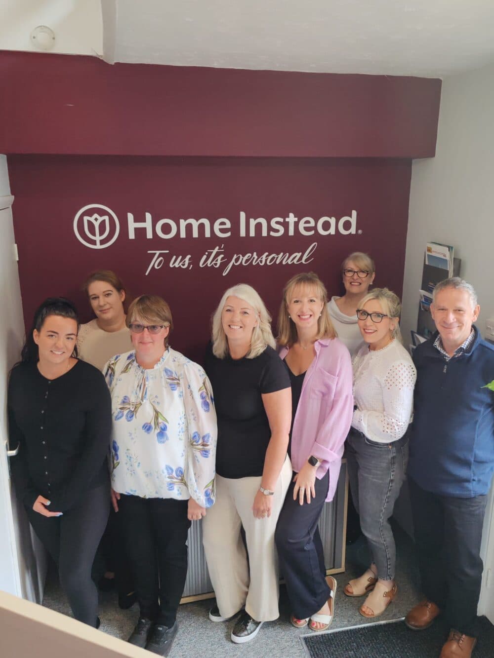 A group of eight people standing and smiling in front of a "Home Instead" sign in an office setting. - Home Instead