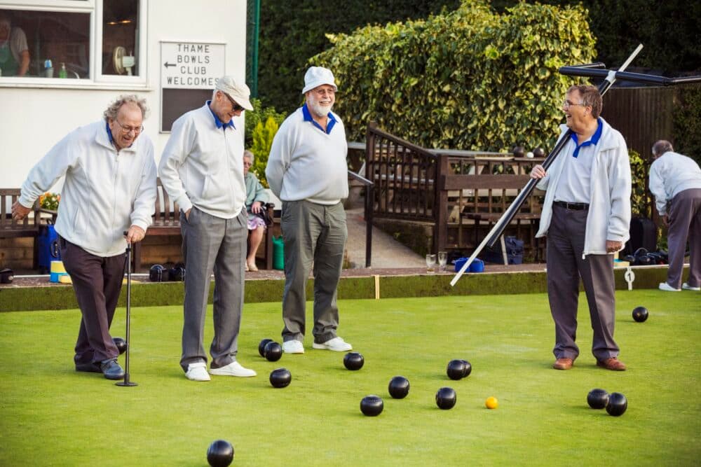 Four elderly men in white uniforms playing lawn bowls, with wooden benches and greenery in the background. - Home Instead