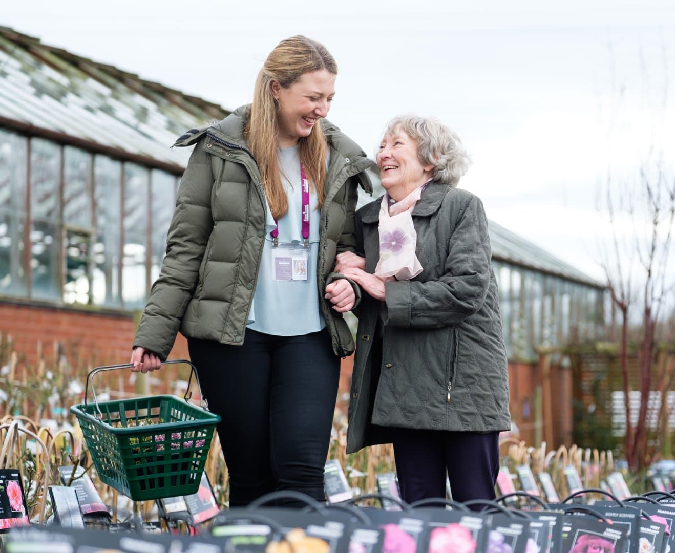 A young woman assists an elderly woman in a garden center, smiling and walking together, carrying a basket. - Home Instead
