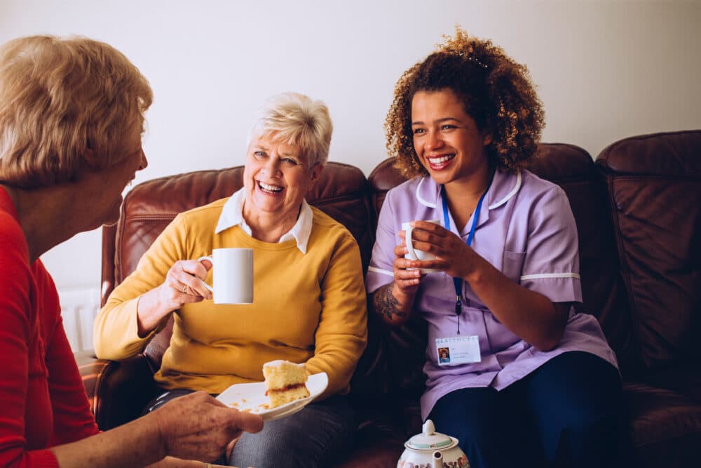 Three women sitting on a couch, smiling and enjoying tea and cake together. One woman is wearing a caregiver uniform. - Home Instead