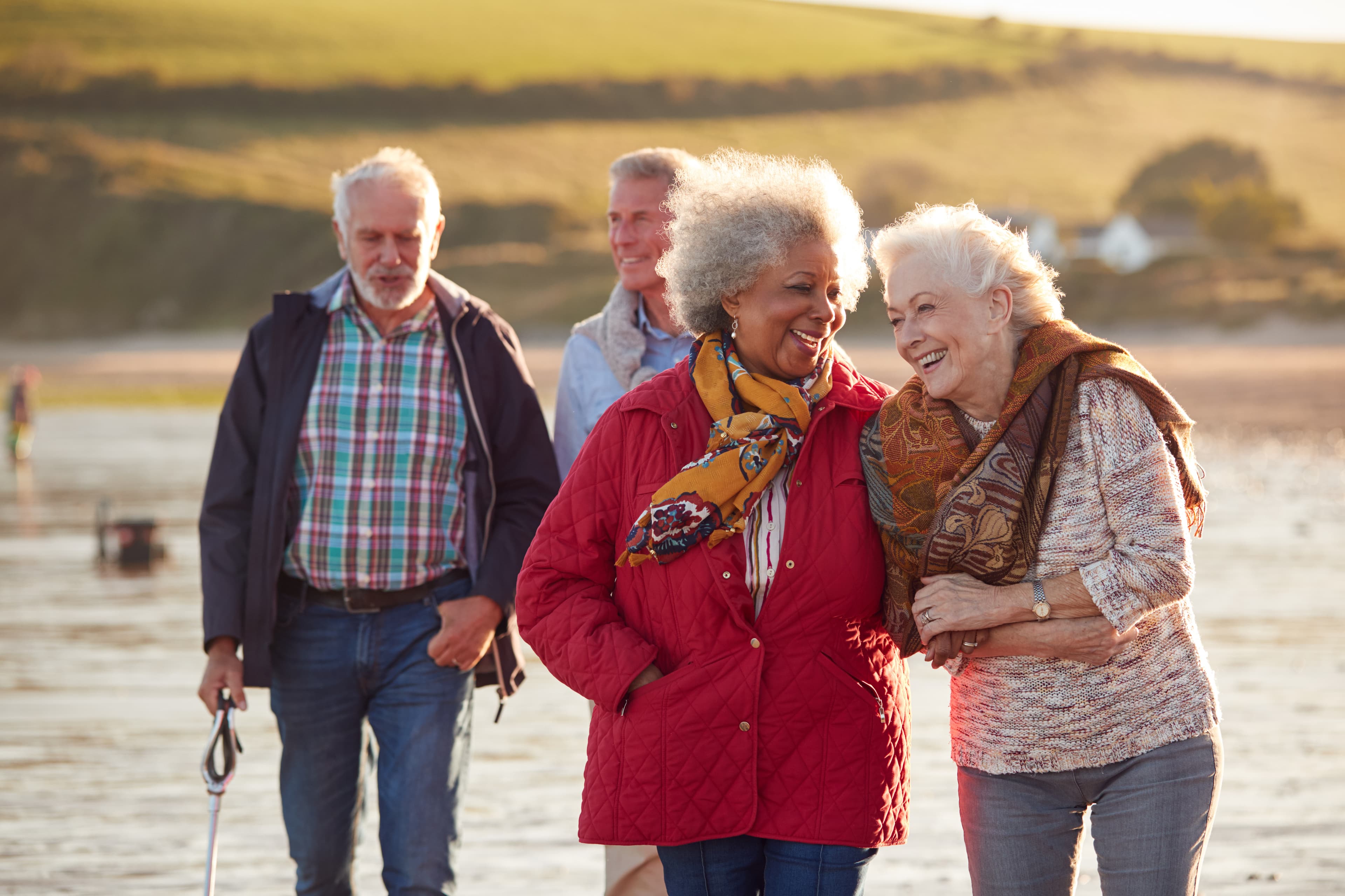 Four elderly friends walking on a beach, two women laughing and the two men walking their dogs in the background. - Home Instead