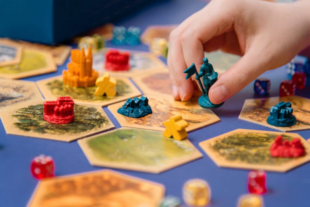 A hand moving a blue knight piece on a board game with hexagonal tiles and various colored dice and pieces. - Home Instead