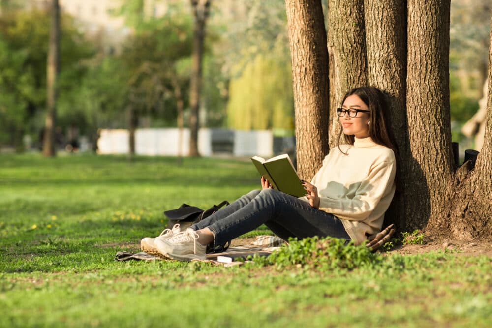 A person wearing glasses sits against a tree reading a book in a sunny park. - Home Instead