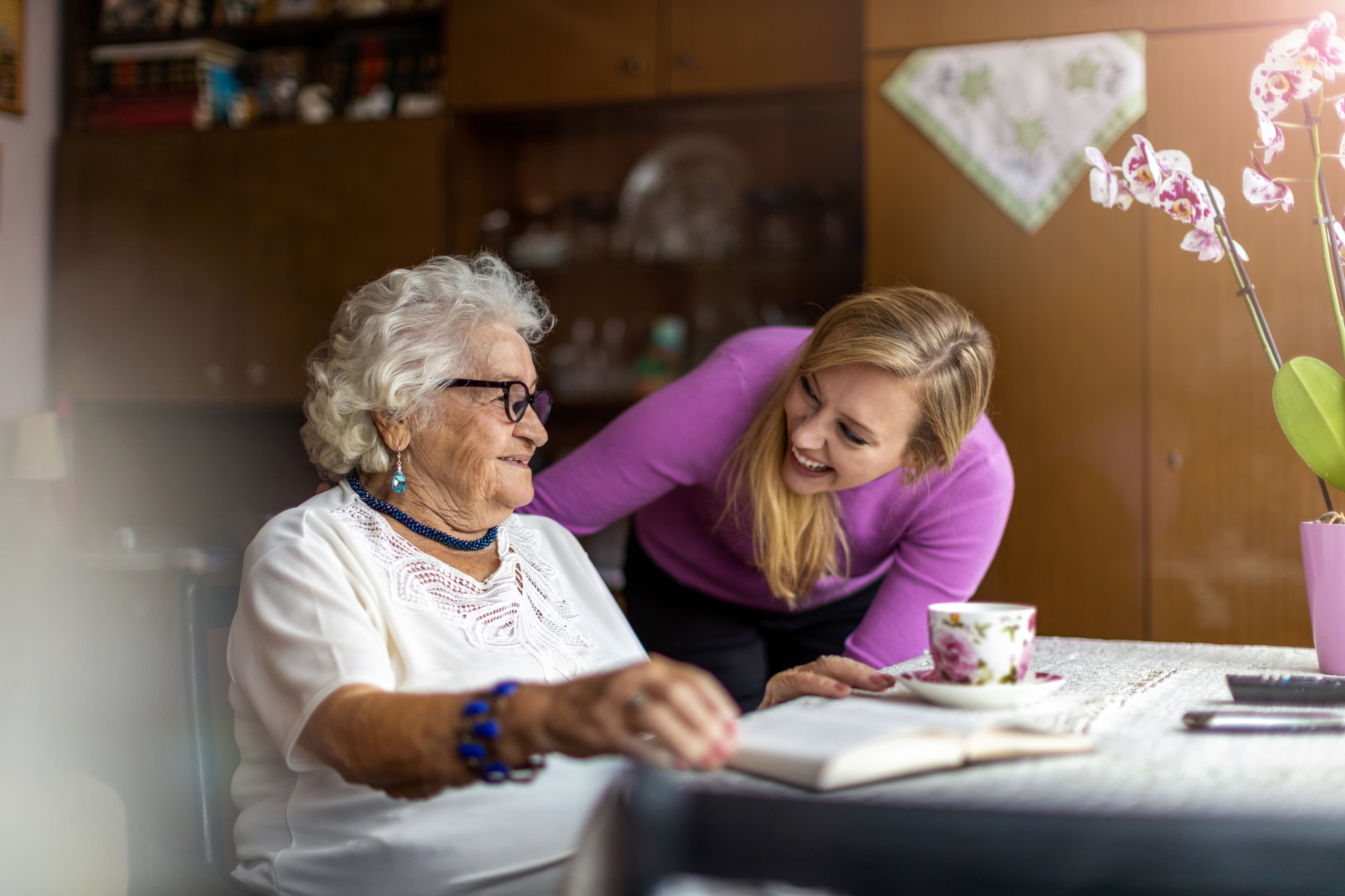 An elderly woman and a younger woman smile and chat over a book at a table with a teacup in a cozy room. - Home Instead