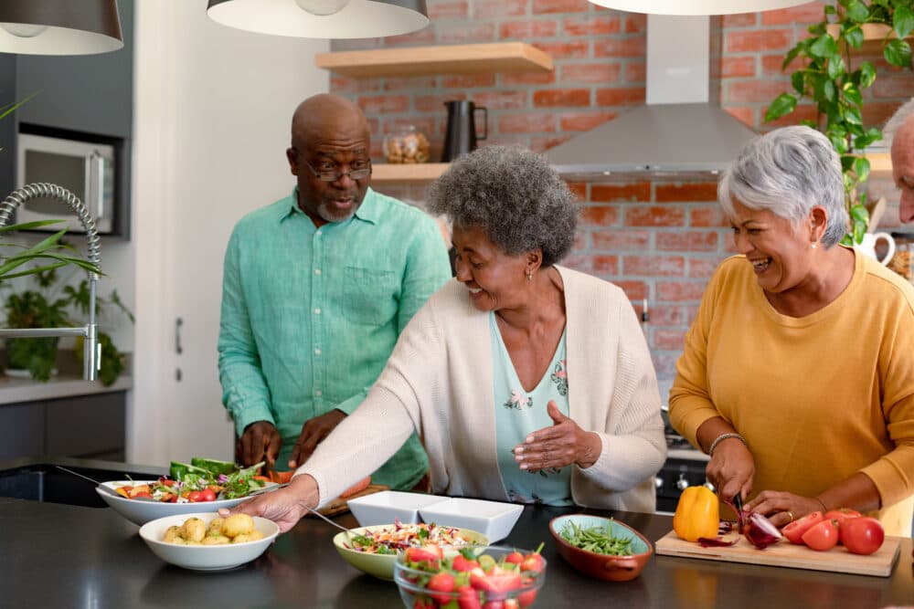 Three elderly friends preparing food together in a modern kitchen, smiling and enjoying their time. - Home Instead
