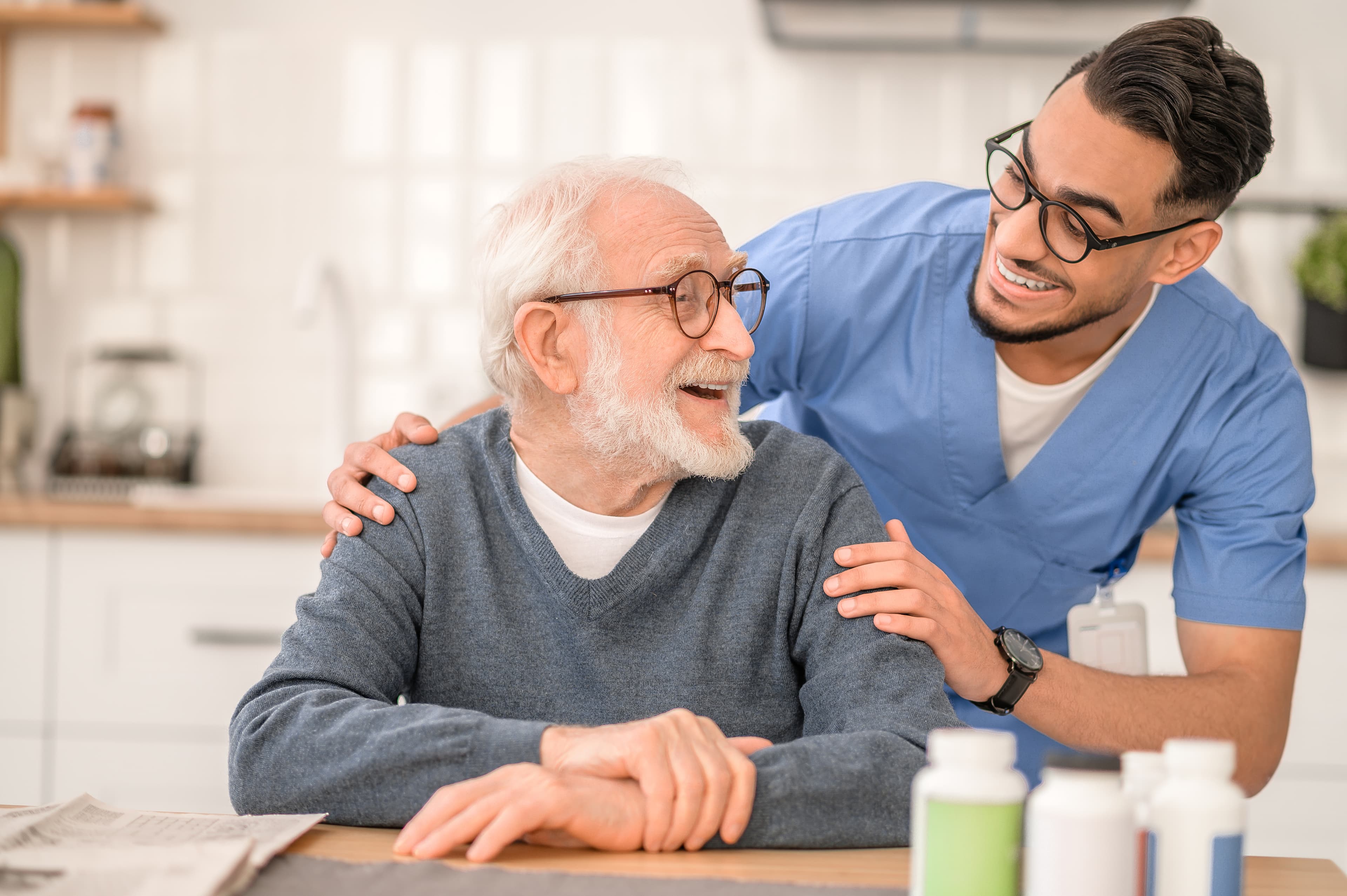 A cheerful elderly man in glasses sits at a table while a smiling young healthcare worker in blue scrubs stands beside him. - Home Instead