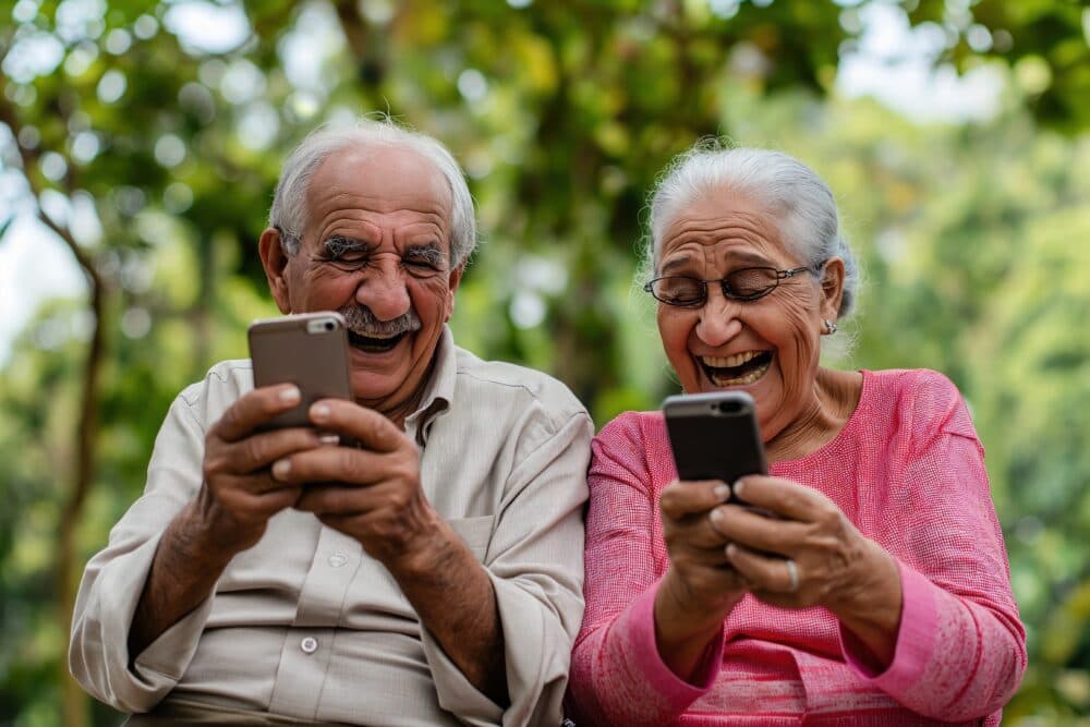 Two senior adults sitting outdoors, laughing and looking at their smartphones. - Home Instead