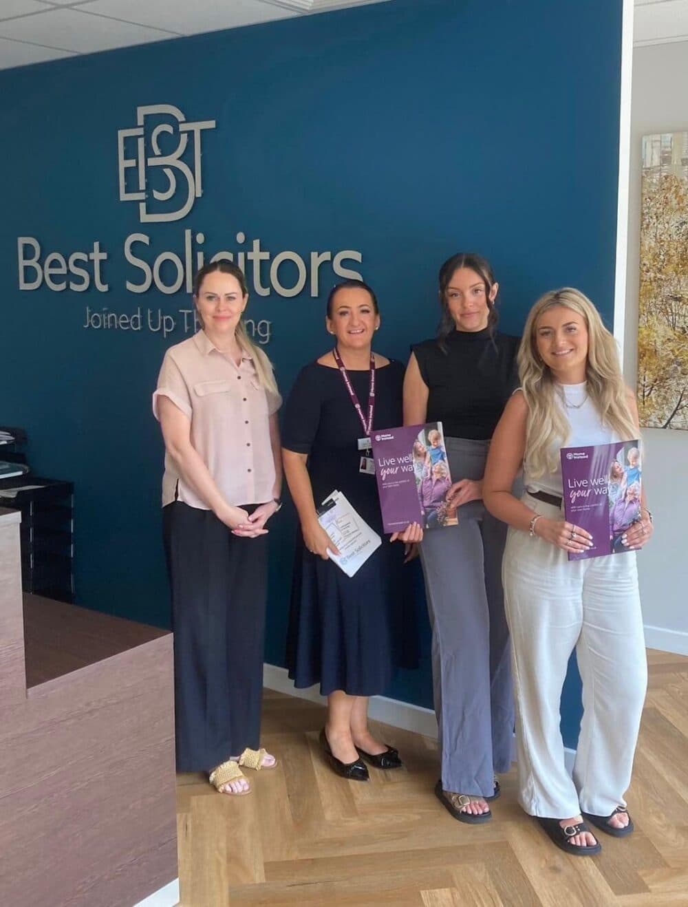 Four women stand smiling in an office, holding brochures, next to a wall sign that reads, "Best Solicitors Joined Up Thinking. - Home Instead
