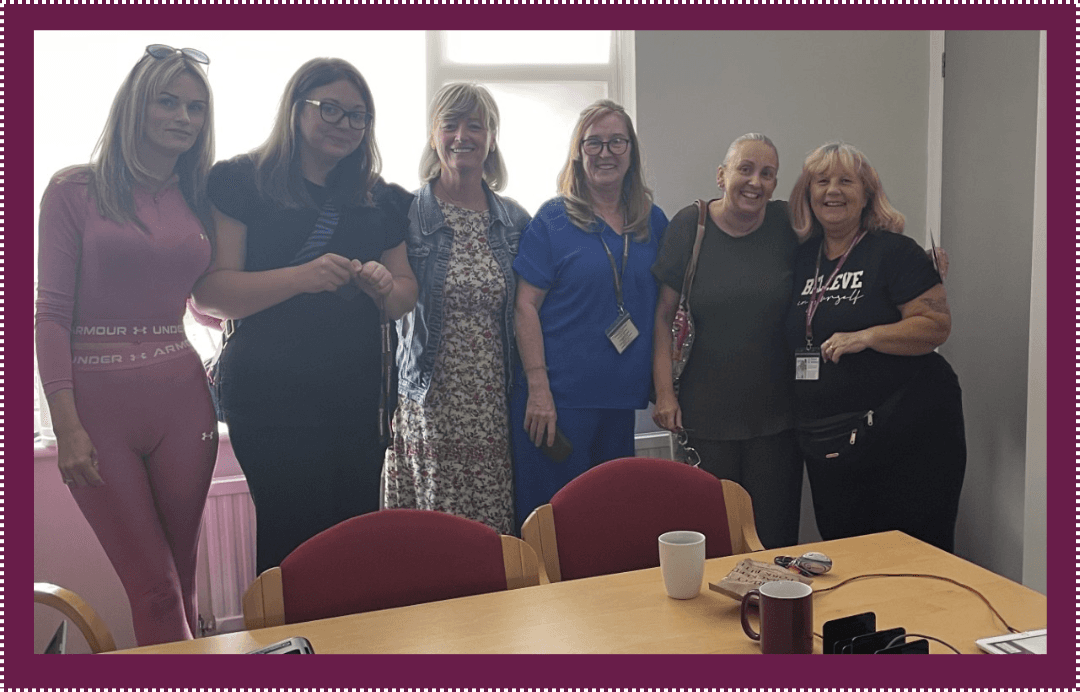 Six women stand close together, smiling, inside an office with a table, mugs, and documents in the foreground. - Home Instead