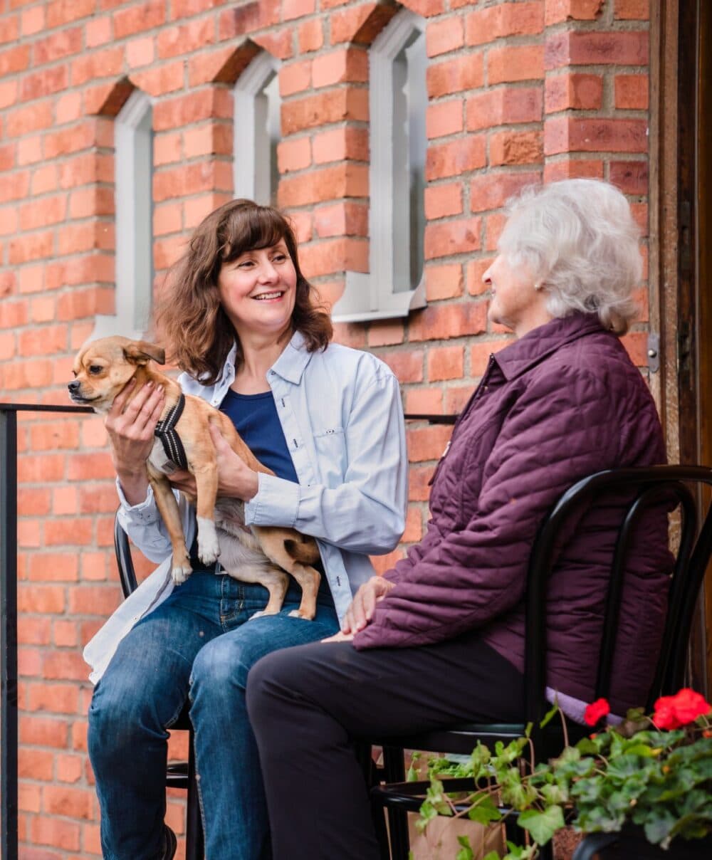 Two women seated outside on stools by a brick wall; the younger woman holding a small dog, both smiling. - Home Instead