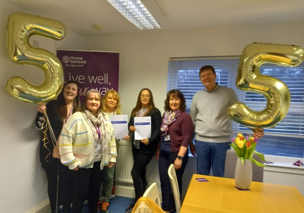 A group of six people holding balloons shaped like the number 5 in an office with a "Home Instead" banner in the background. - Home Instead