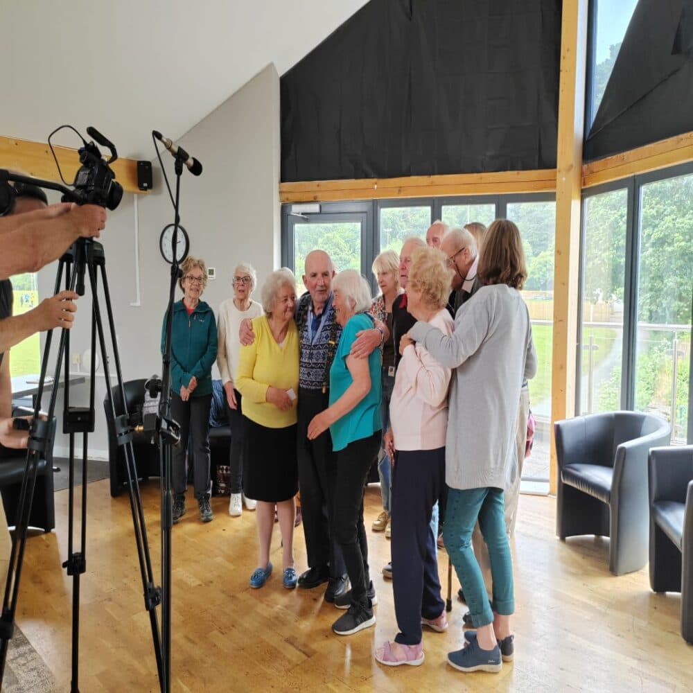 A group of older adults stand together in a room, posing for a camera setup with microphones. - Home Instead