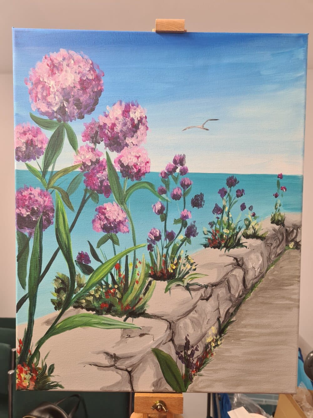 A painting of pink and purple flowers by the seaside, with a stone path and a seagull flying in the sky