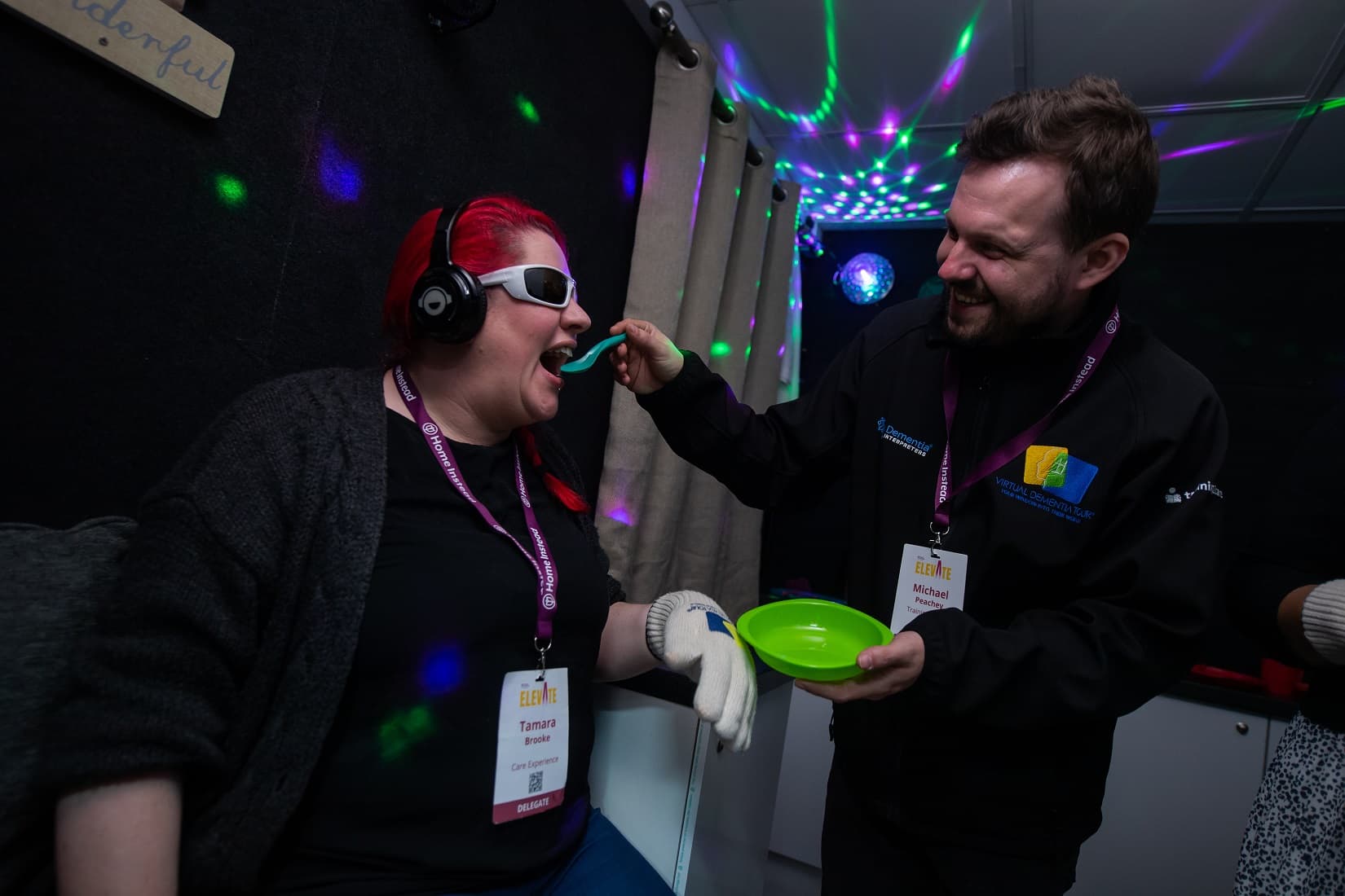 A person in a black shirt is feeding a laughing person wearing headphones and sunglasses under colorful lights. - Home Instead
