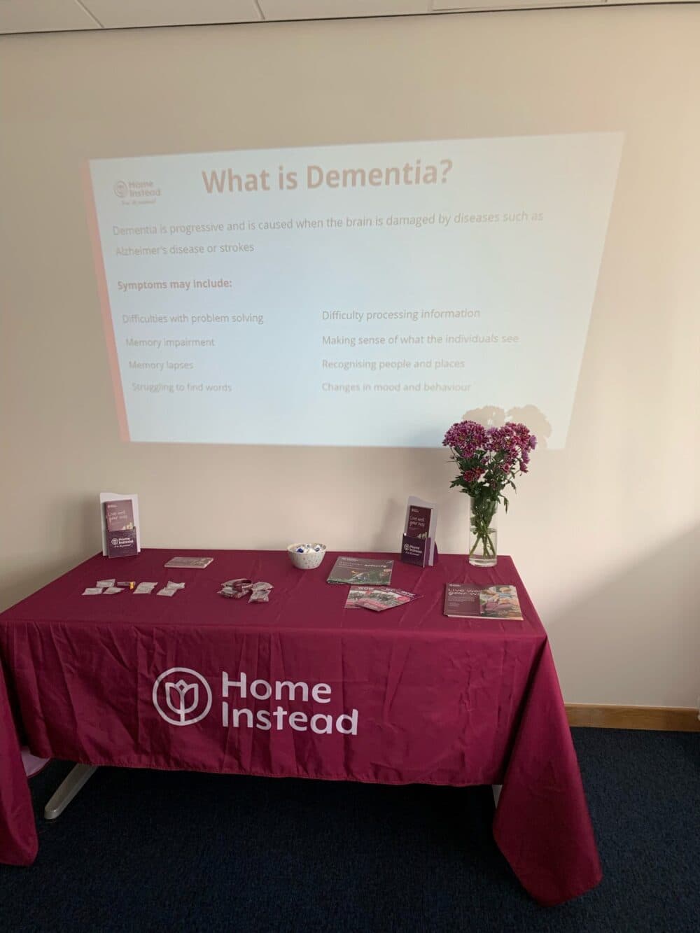A table with a purple tablecloth featuring Home Instead and dementia awareness materials, with a projection on the wall. - Home Instead