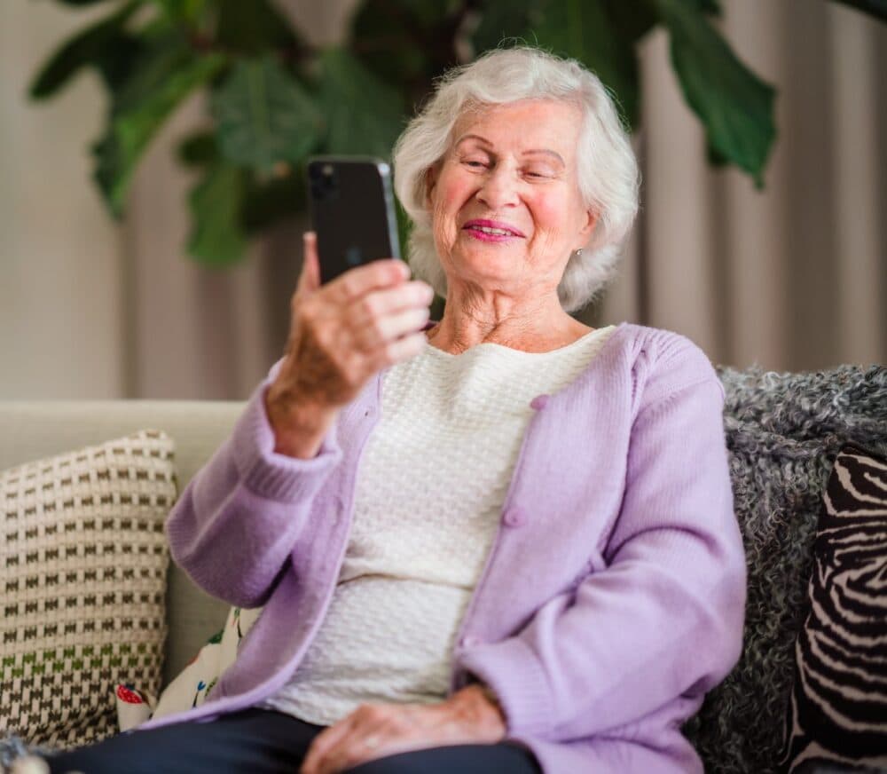 Elderly woman with white hair, smiling and holding a smartphone while sitting on a couch. - Home Instead