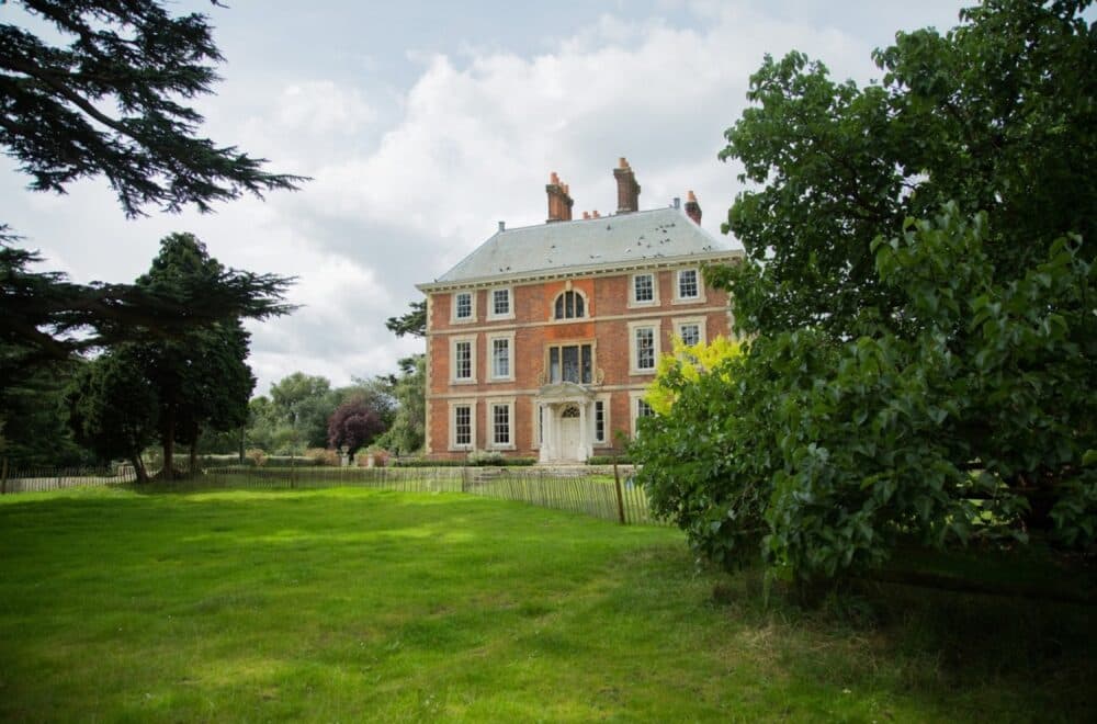 A three-story brick mansion with a large lawn and trees in the foreground on a partly cloudy day. - Home Instead
