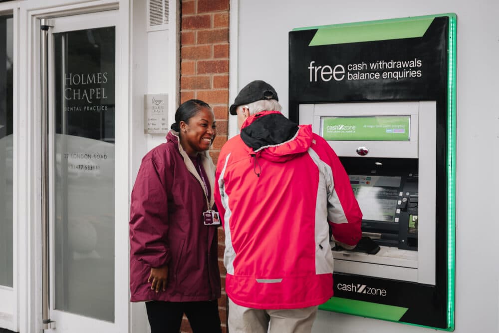 A woman and a man are using an outdoor ATM. The woman is smiling while the man operates the machine. - Home Instead