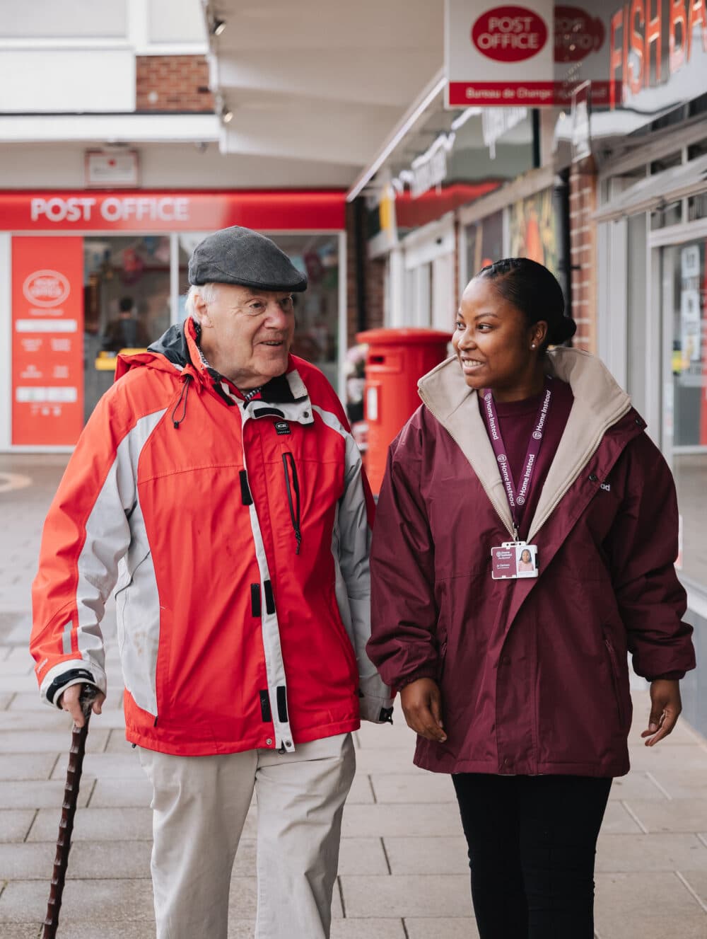 An elderly man with a cane walks beside a younger woman outside a post office. Both are wearing jackets and smiling. - Home Instead
