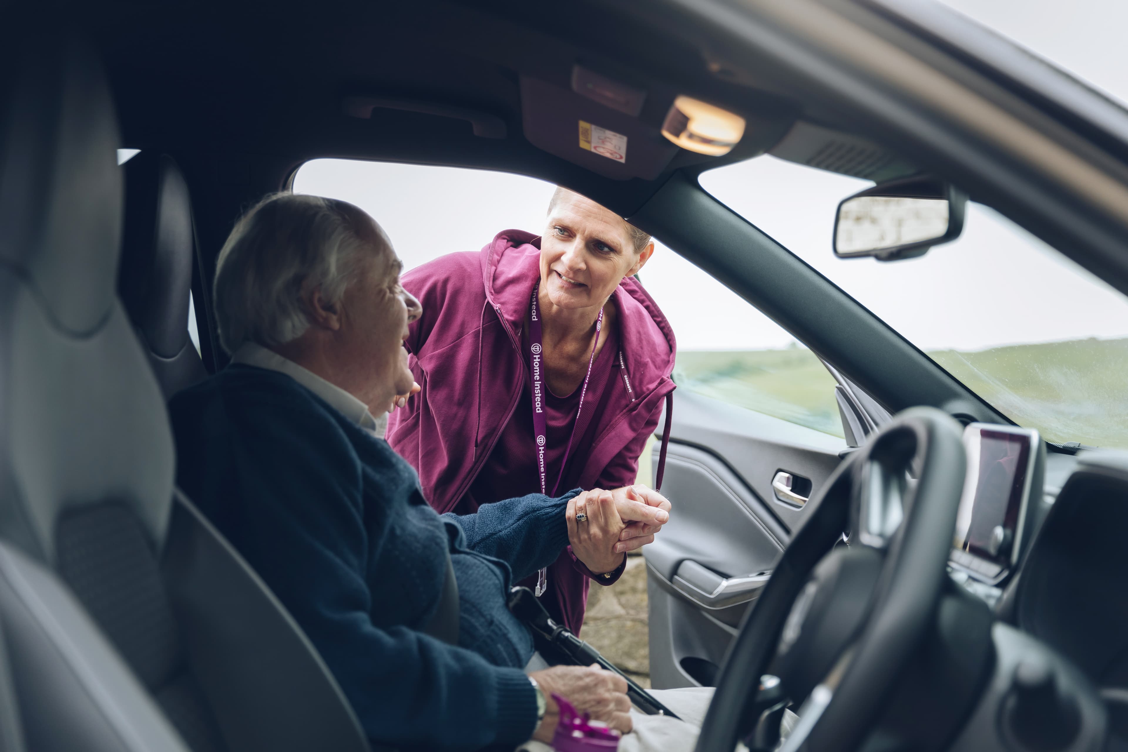 A caregiver assists an elderly person sitting in a car, holding hands and smiling warmly. - Home Instead