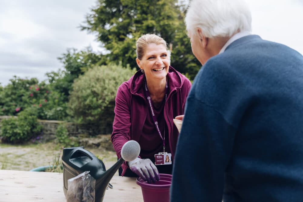 A smiling woman in a purple jacket helps an elderly person with gardening outdoors, holding a purple pot. - Home Instead